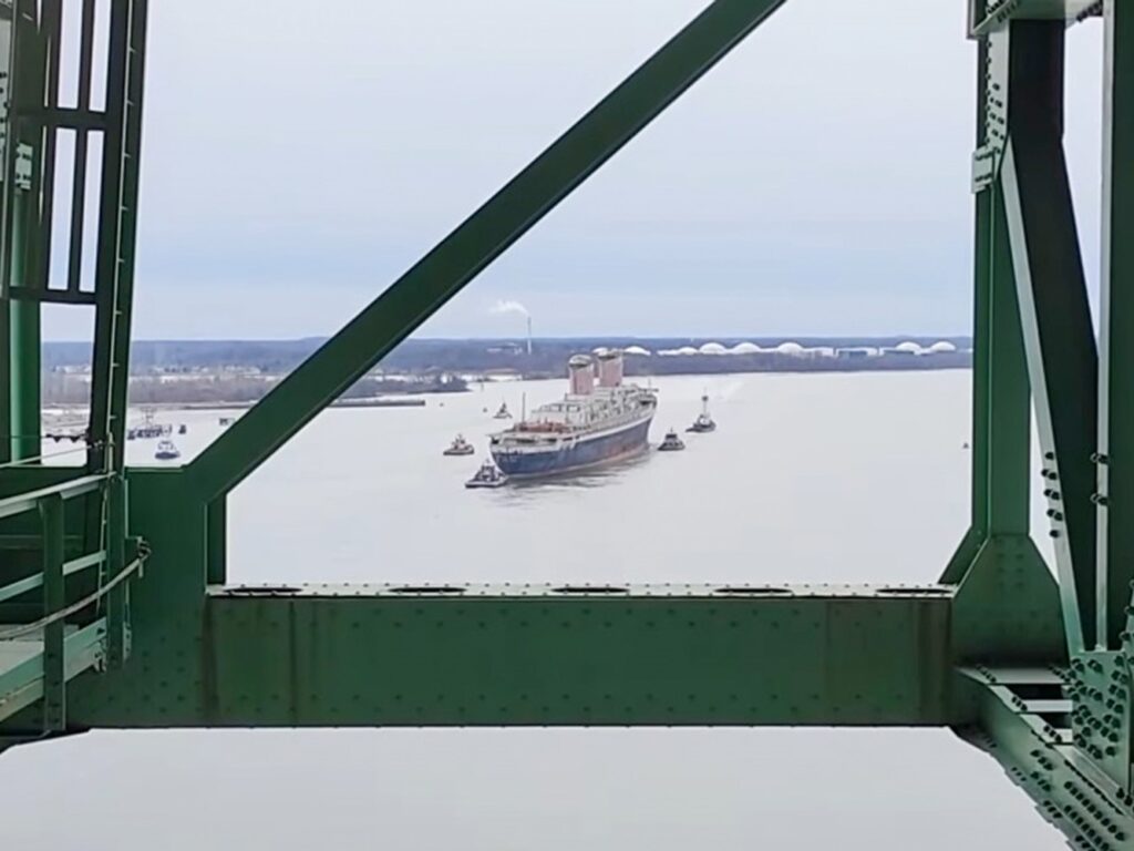 An old ocean liner sails down a river assisted by tugboats.