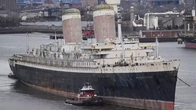 An old ocean liner sails down a river assisted by tugboats.
