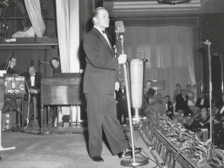 A black and white image of a man in a tuxedo singing for a crowd.