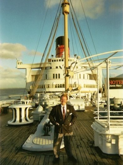 A man in a blue blazer and grey pants standing on a ship's deck. A red and black funnel is seen in the background.