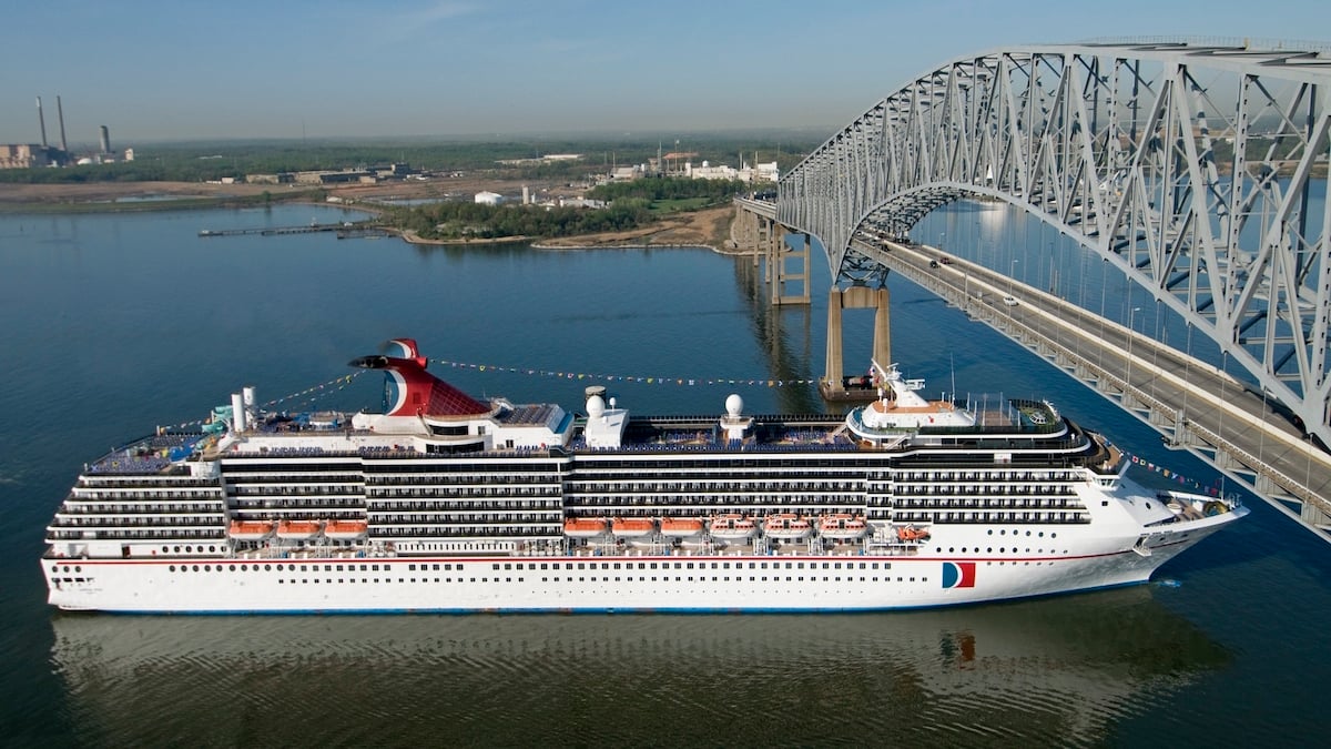 A white cruise ship with a red, white, and blue funnel sails under a steel frame bridge.