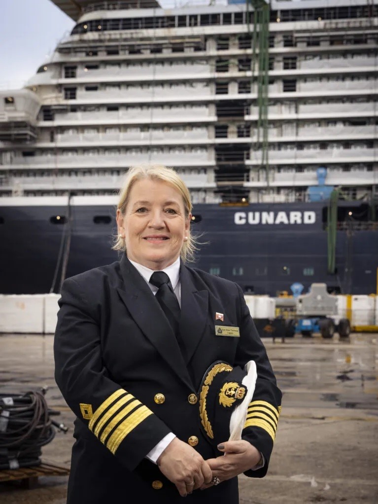 A woman in a blue uniform and gold braid on her sleeves stands in front of a ship.