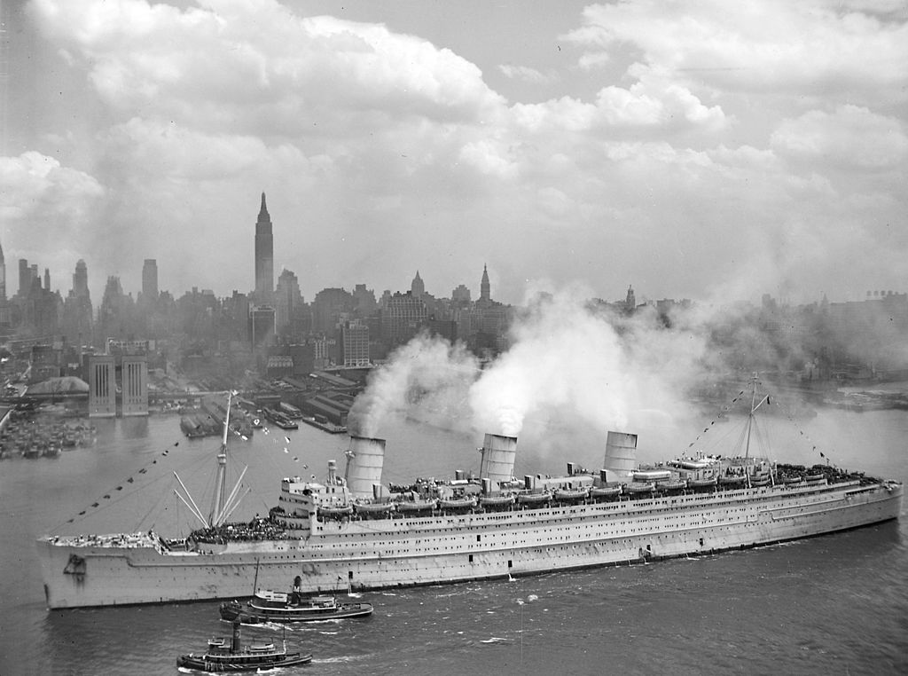 A black and white image of a large ocean liner sailing past New York City.