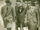 A black and white image of men in hats and trench coats walking together.
