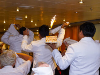 A group of waiters in white jackets carry a dessert with a flaming candle on it.
