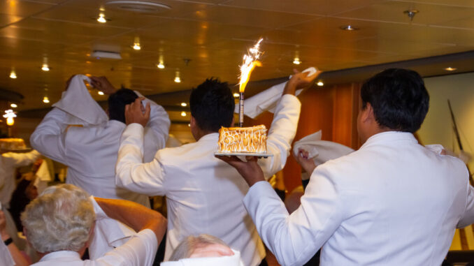 A group of waiters in white jackets carry a dessert with a flaming candle on it.