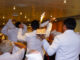A group of waiters in white jackets carry a dessert with a flaming candle on it.