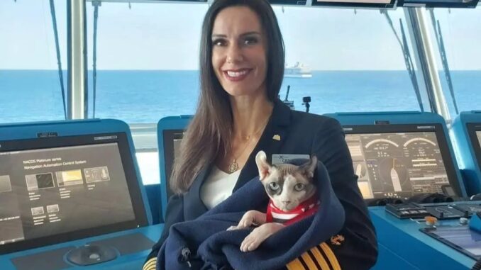 A woman in a ship's captain uniform holding a small hairless cat wrapped up in a blue blanket.