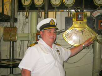 A man in a white uniform smiles while pointing at a brass engine order telegraph.