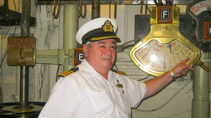 A man in a white uniform smiles while pointing at a brass engine order telegraph.