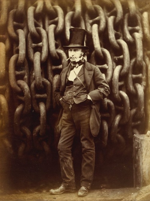 A dapper man in a suit and hat stands in front of giant chains.