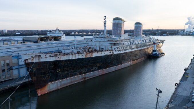 A rusty, decrepit ocean liner sits at a pier in the morning light.