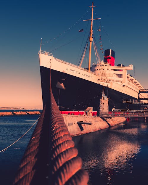 A giant ocean liner tied up to a pier. It has a black hull and red funnel.