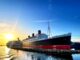 A black and red ocean liner at dock with the sun rising behind it.